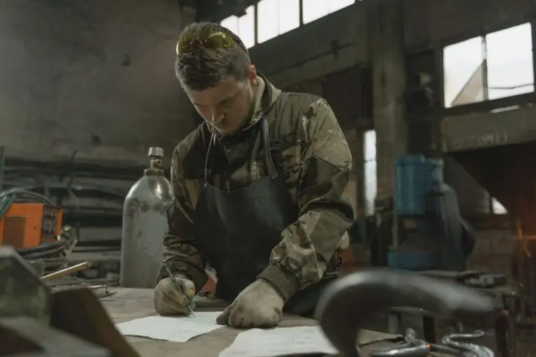 A welder at a shop bench writing on paper, the bookkeeping side of 1099 vs W-2 work for a traveling tradesperson